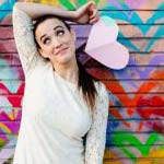 Magician Kayla Drescher stands in front colorful graffiti holding a paper cut-out heart